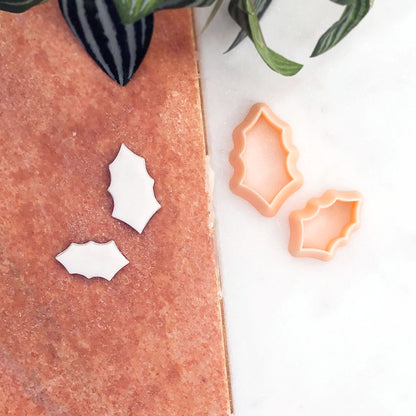 Two pale peach cookie cutters shaped like holly leaves, shown beside a copper-toned board with two white cutout pieces on a white surface, with green leaves at the top edge.