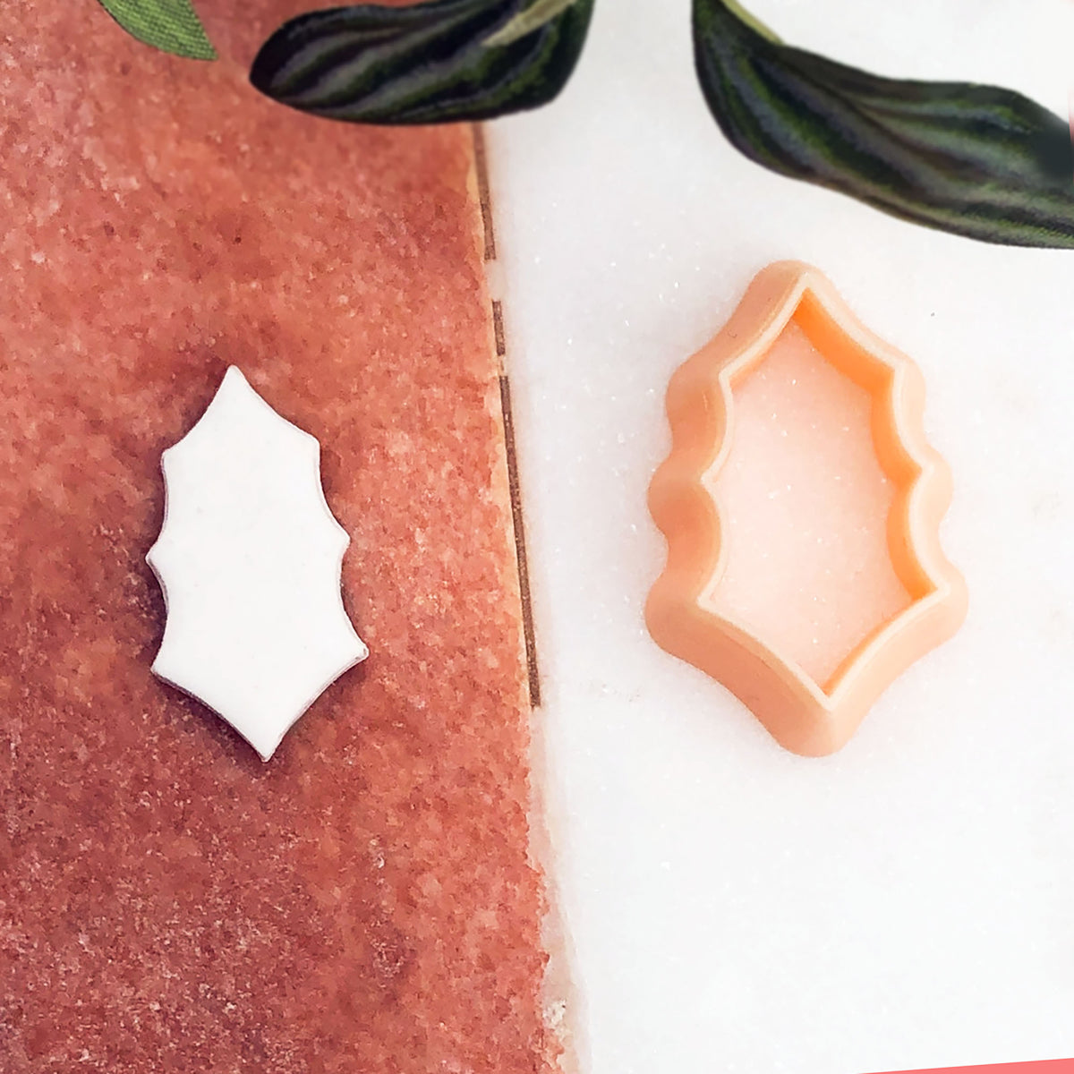 A cookie cutter in peach-colored silicone sits on a white surface next to a white holly leaf cut out of the shape, with a pinkish-brown textured surface on the left and green leaves at the top.