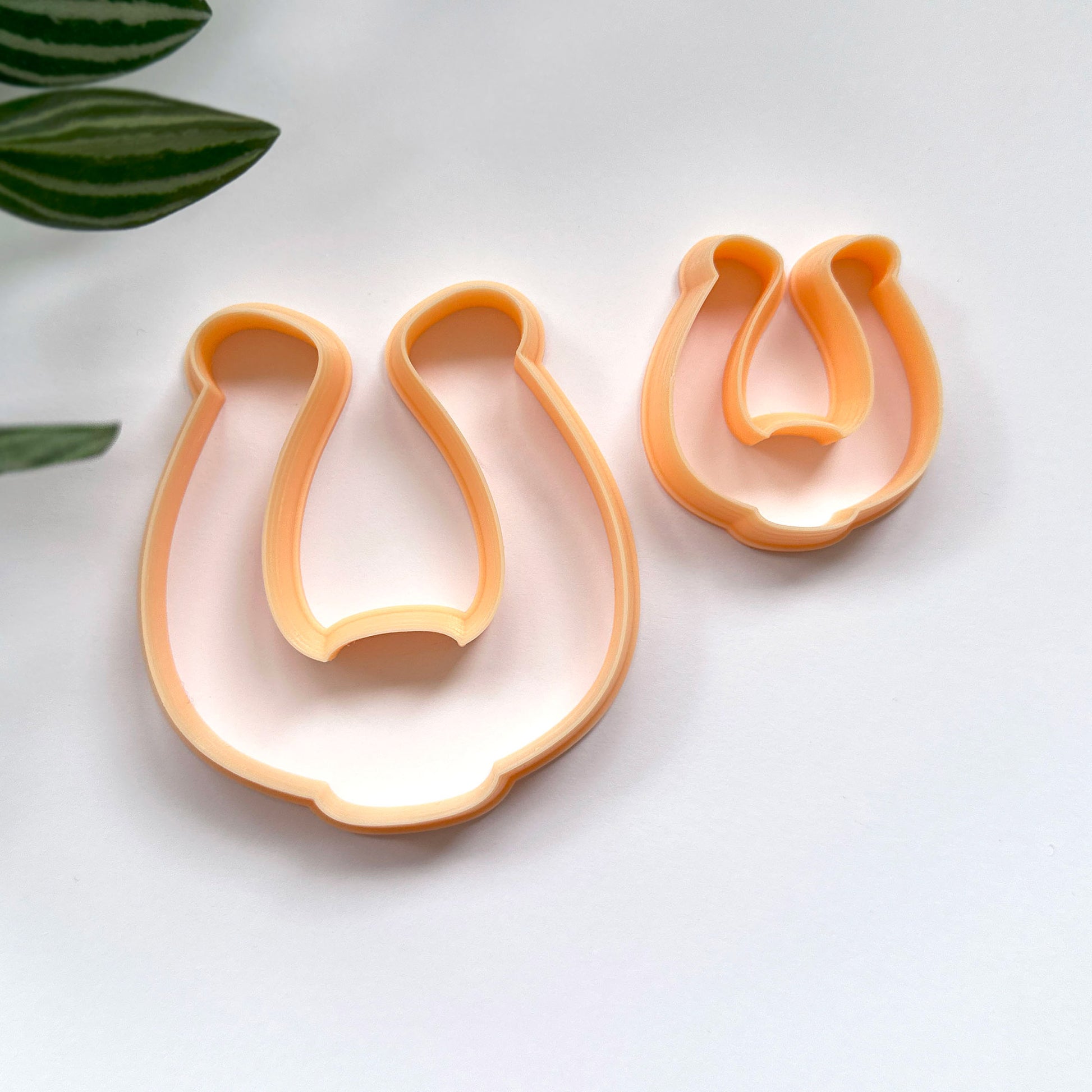 Two orange horseshoe shaped cookie cutters, one large and one small, displayed on a clean white surface with a green plant leaf in the corner.