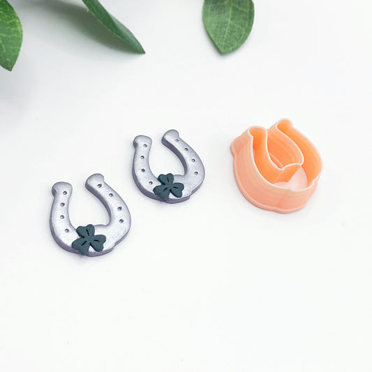 Two small silver horseshoe-shaped polymer clay cut outs with dark green clover details are shown beside a peach-colored clay cutter on a white background with a few green leaves at the top.