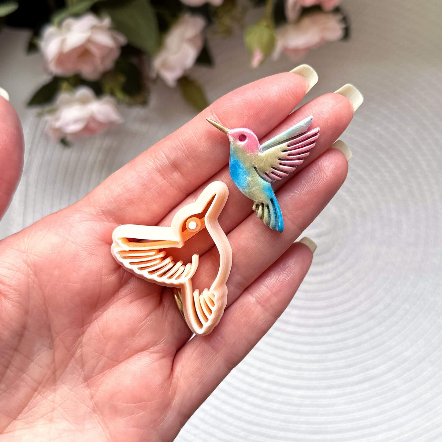 Hummingbird clay cutter with the polymer clay cut out on a textured surface with pink flowers in the background.