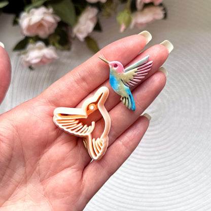 Hummingbird clay cutter with the polymer clay cut out on a textured surface with pink flowers in the background.