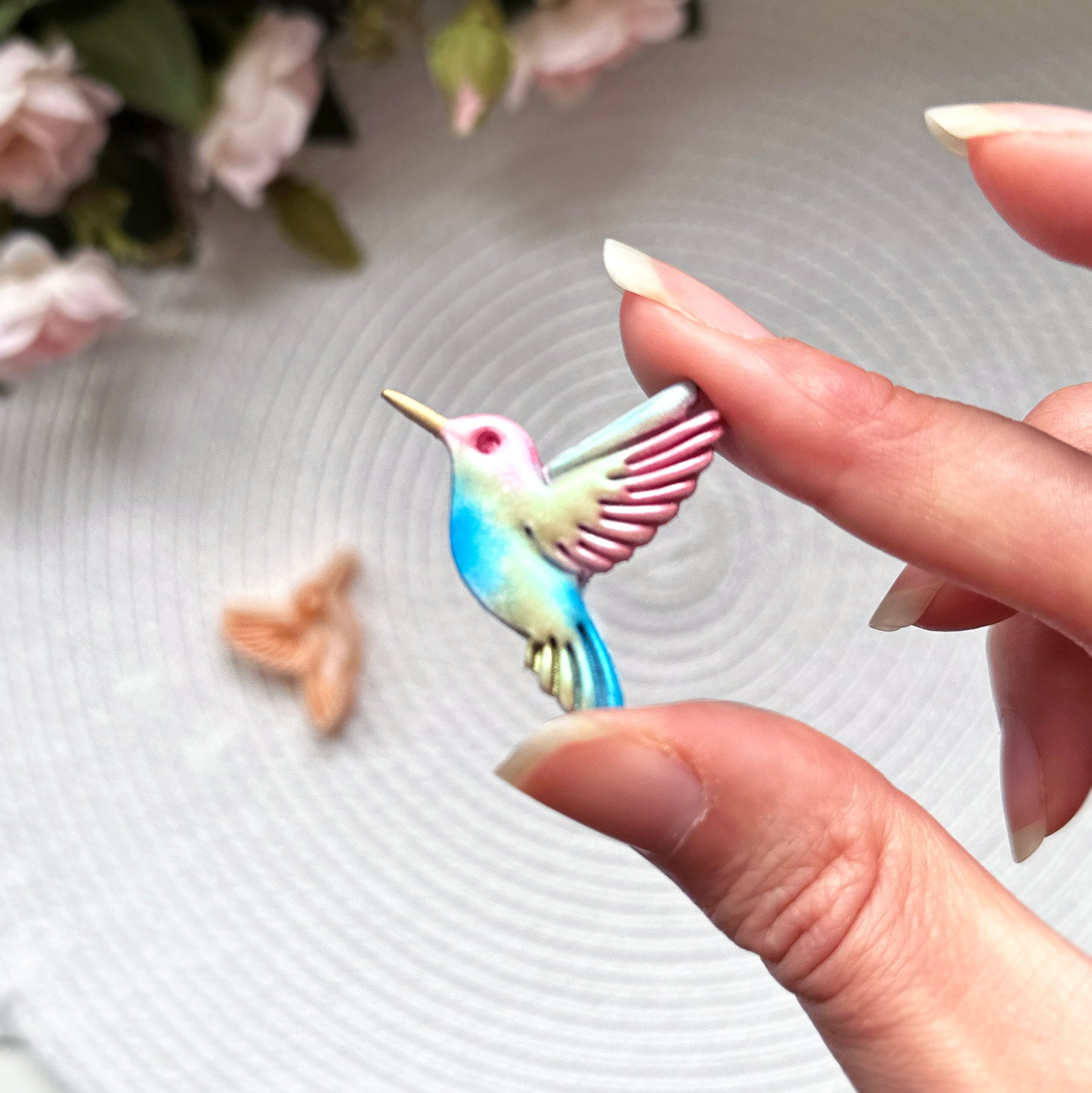 Hummingbird clay cutter with the polymer clay cut out on a textured surface with pink flowers in the background.