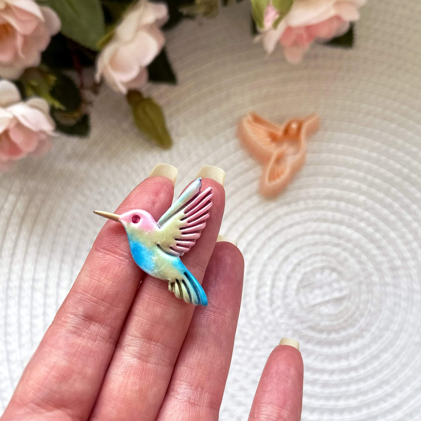 Hummingbird clay cutter with the polymer clay cut out on a textured surface with pink flowers in the background.