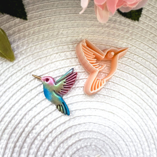 Hummingbird clay cutter with the polymer clay cut out on a textured surface with pink flowers in the background.