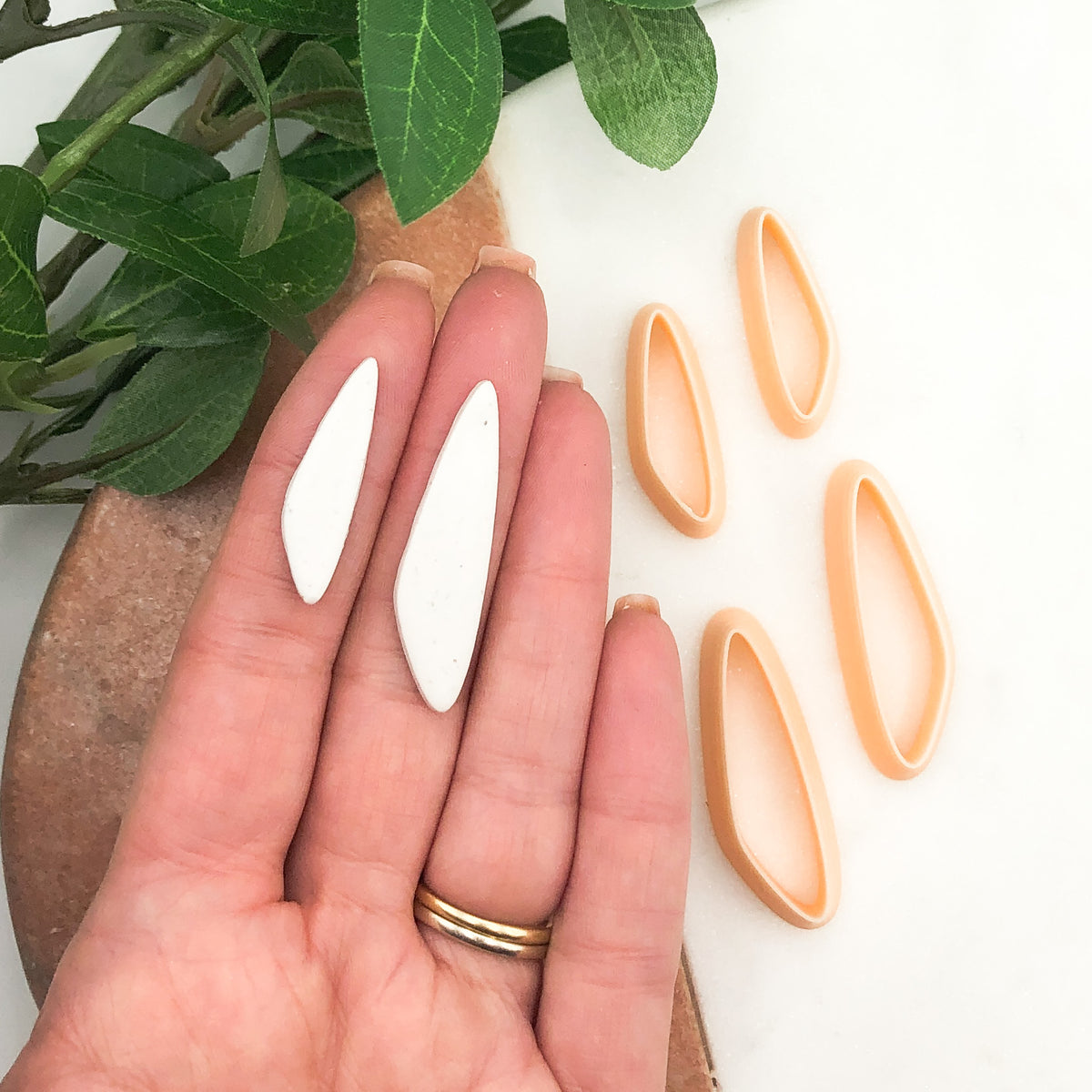 organic slim drop clay cutter, shown as a mirror set. Two clay cut outs on a hand then two mirroed peach clay cutters on a marble tray with green leaves in the background.