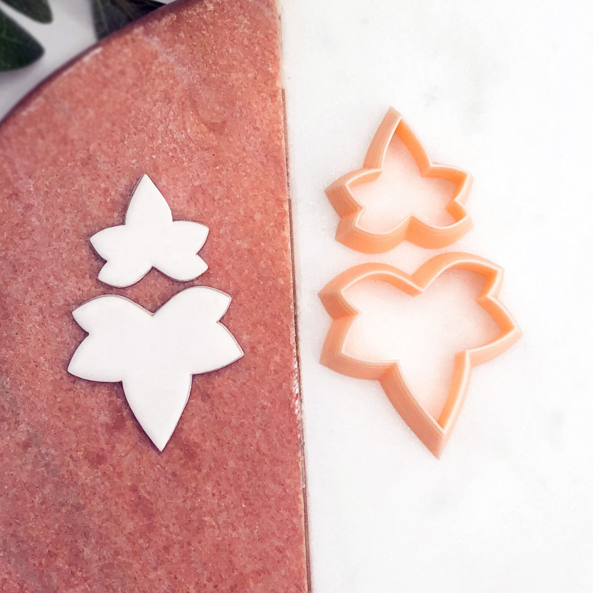 Two pale peach ivy leaf-shaped cookie clay cutters on a white marble surface next to matching white cutouts on a dusty rose board.