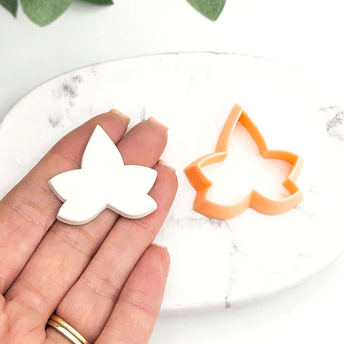 A hand holds a white clay ivy leaf-shaped piece next to an orange plastic leaf-shaped cookie clay cutter on a white marble surface.