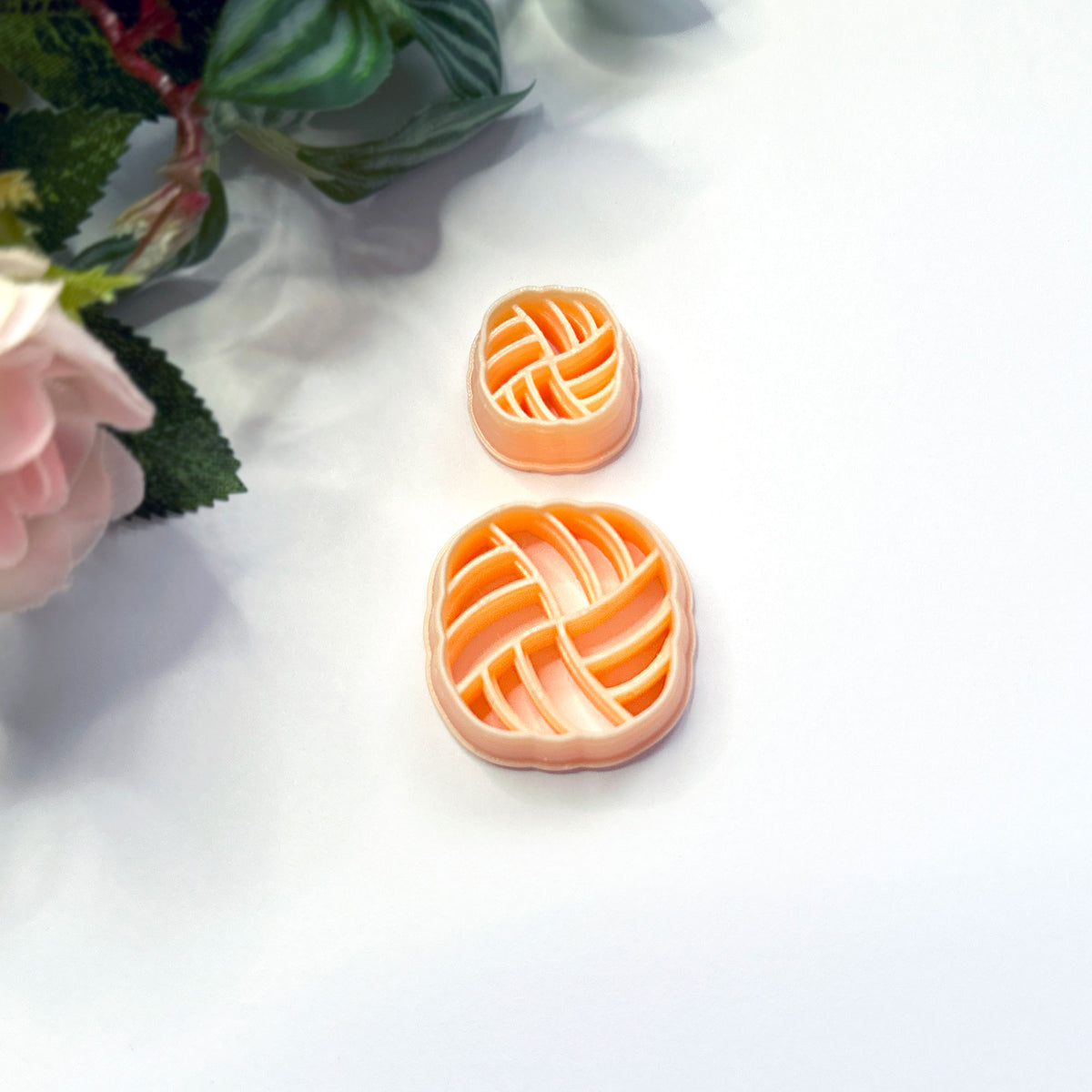 Two peach-colored plastic PLA clay cookie cutters with a braided or woven pattern, displayed on a white surface with decorative pink flowers in the corner.