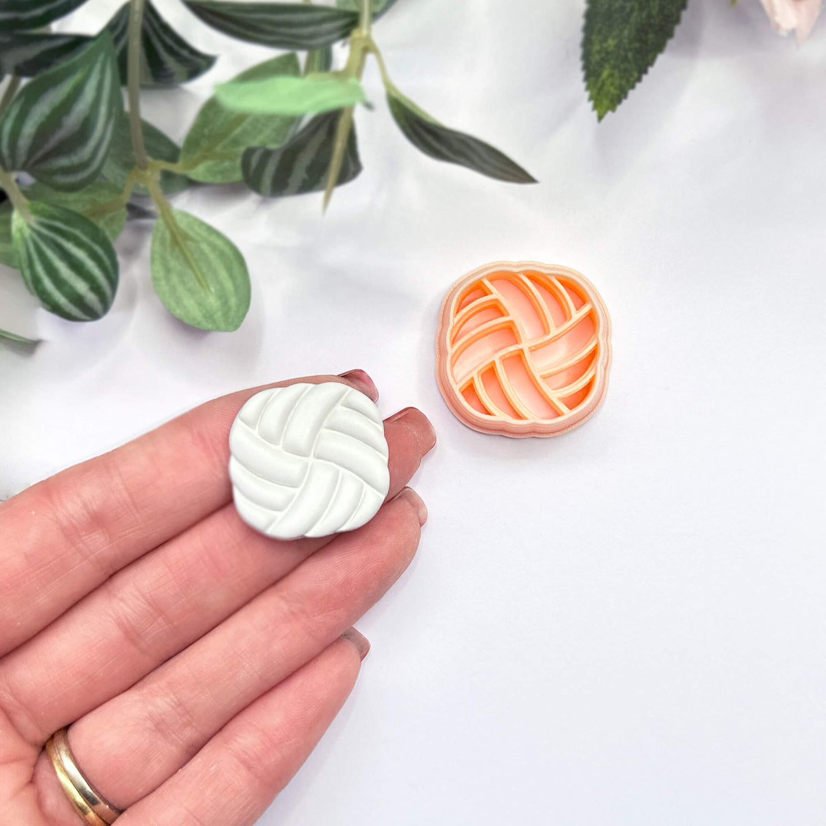 A hand holds a small white, rounded-square polymer clay cutout with a braided or woven pattern, while a matching peach cutter sits on a white surface nearby. Green leaves are softly blurred in the background.