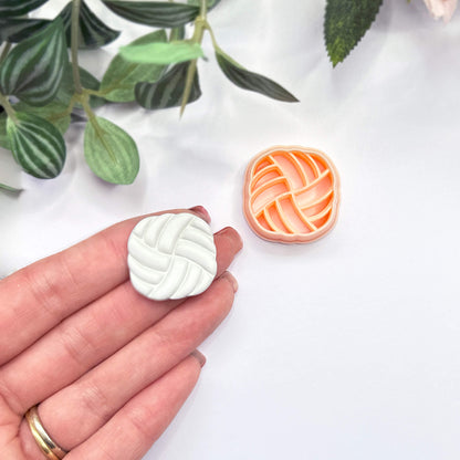 A hand holds a small white, rounded-square polymer clay cutout with a braided or woven pattern, while a matching peach cutter sits on a white surface nearby. Green leaves are softly blurred in the background.