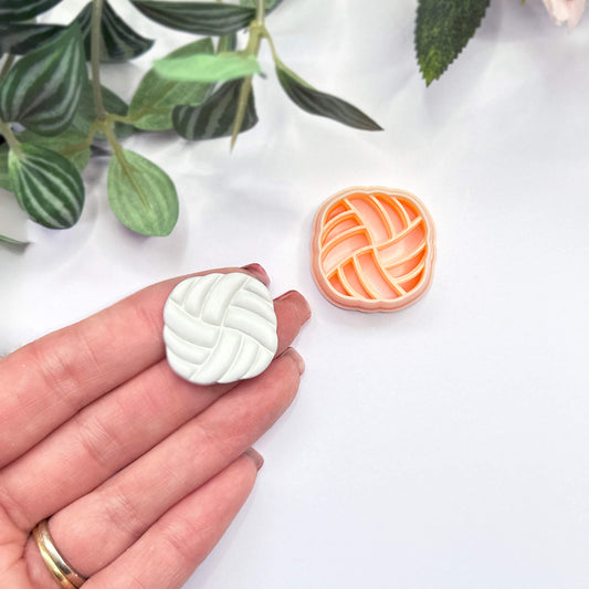 A hand holds a small white, rounded-square polymer clay cutout with a braided or woven pattern, while a matching peach cutter sits on a white surface nearby. Green leaves are softly blurred in the background.