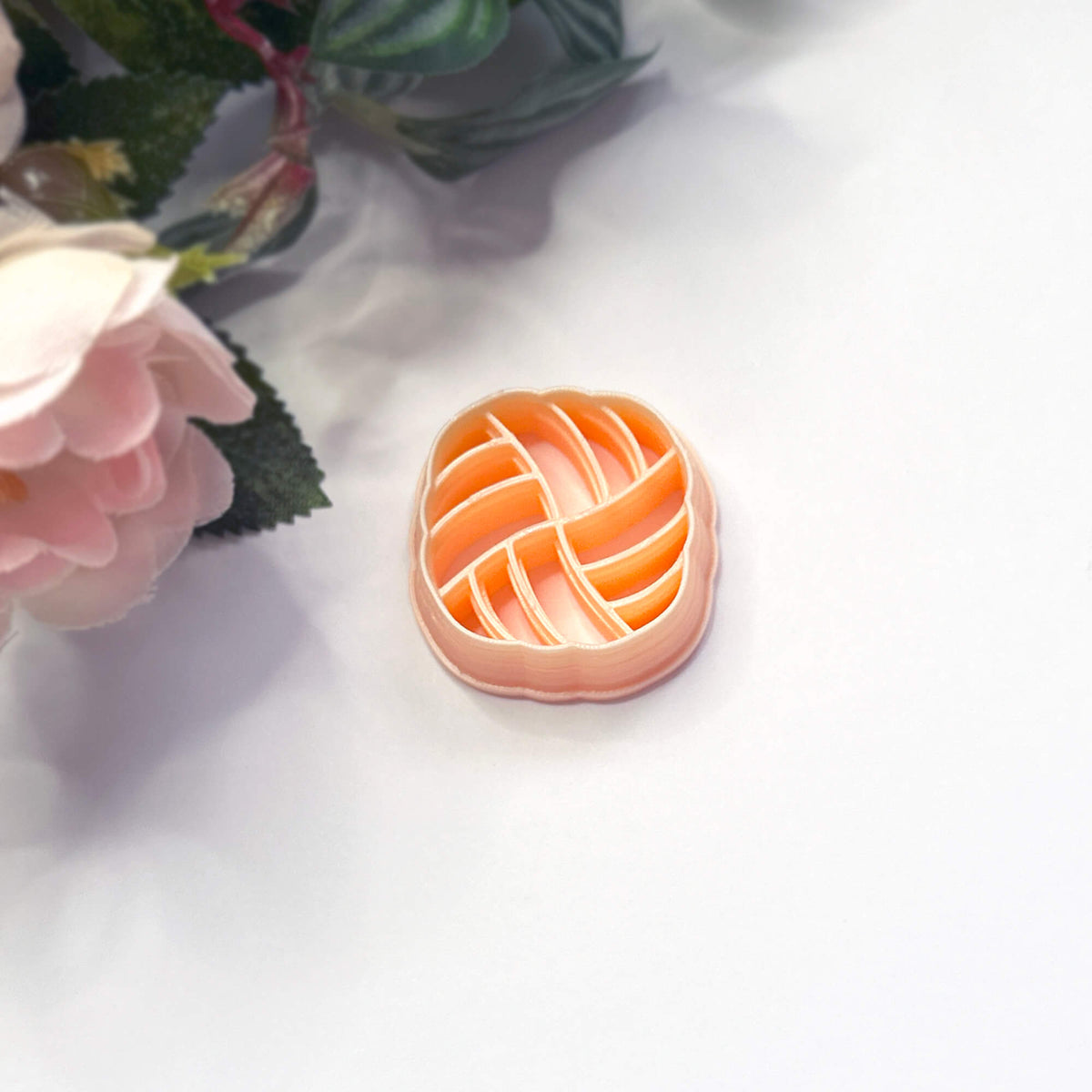 A small peach polymer clay cutter in a   striped basketweave pattern, placed on a white surface with pink flowers in the background.