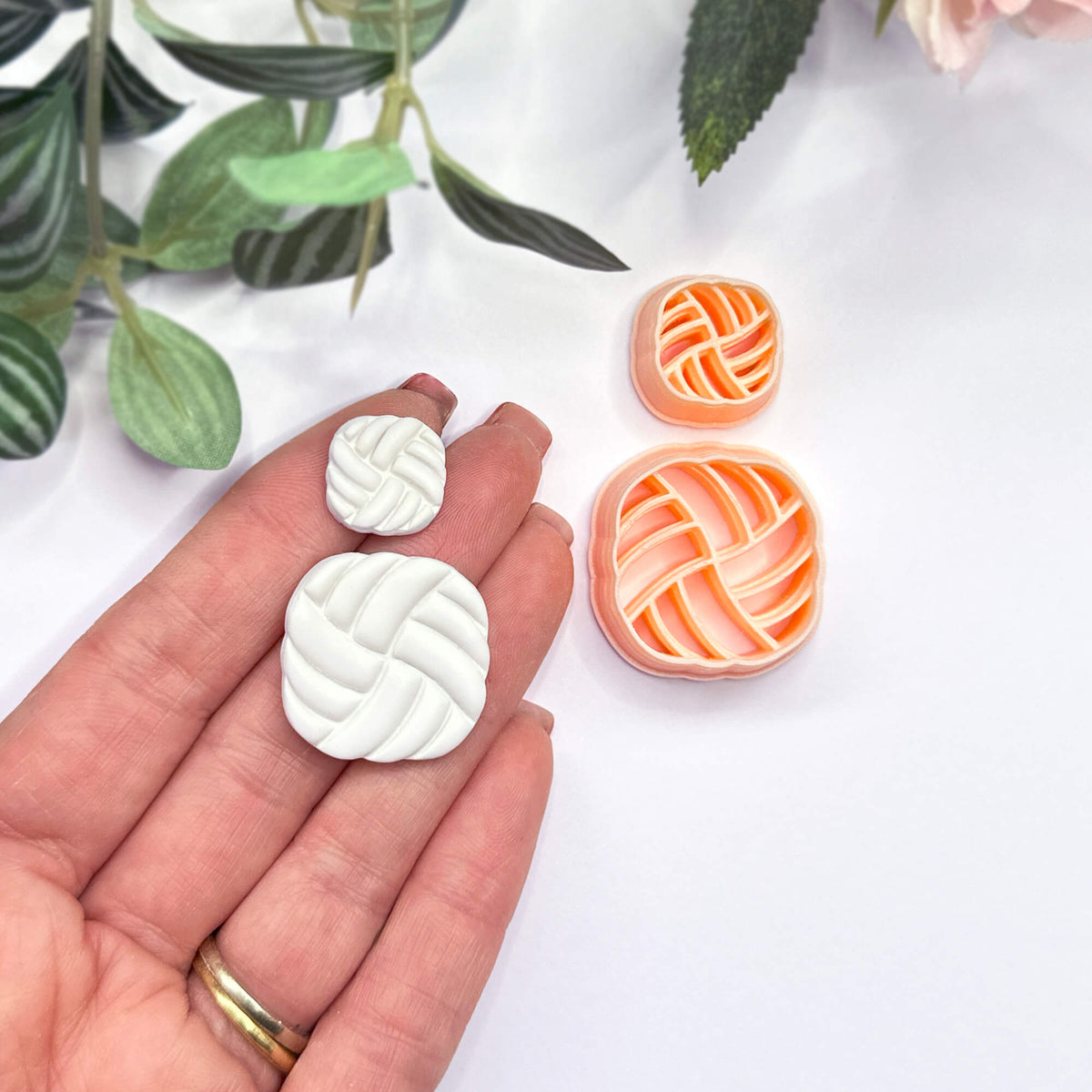 A hand holds two small white, rounded-square polymer clay cutouts with a braided or woven pattern, while the matching peach cutters sit on a white surface nearby. Green leaves are softly blurred in the background.