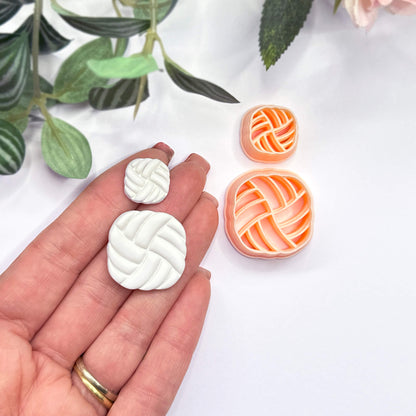 A hand holds two small white, rounded-square polymer clay cutouts with a braided or woven pattern, while the matching peach cutters sit on a white surface nearby. Green leaves are softly blurred in the background.