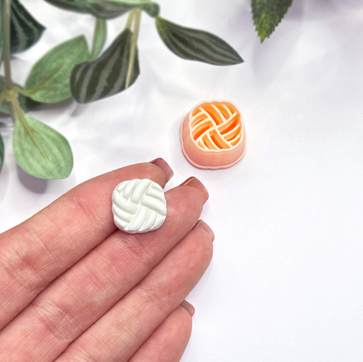 A hand holds a small white, rounded-square polymer clay cutout with a braided or woven pattern, while a matching peach cutter sits on a white surface nearby. Green leaves are softly blurred in the background.
