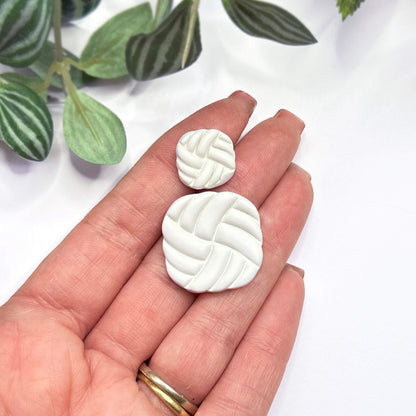 Two small white textured clay charms shaped like woven squares, held in a person's hand against a white background with green leaves in the corner.