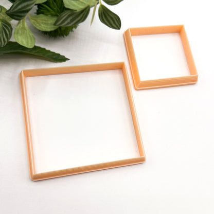 Two square plastic cookie cutters in a light peach-orange color, shown on a white surface with green leaves in the top-left corner.
