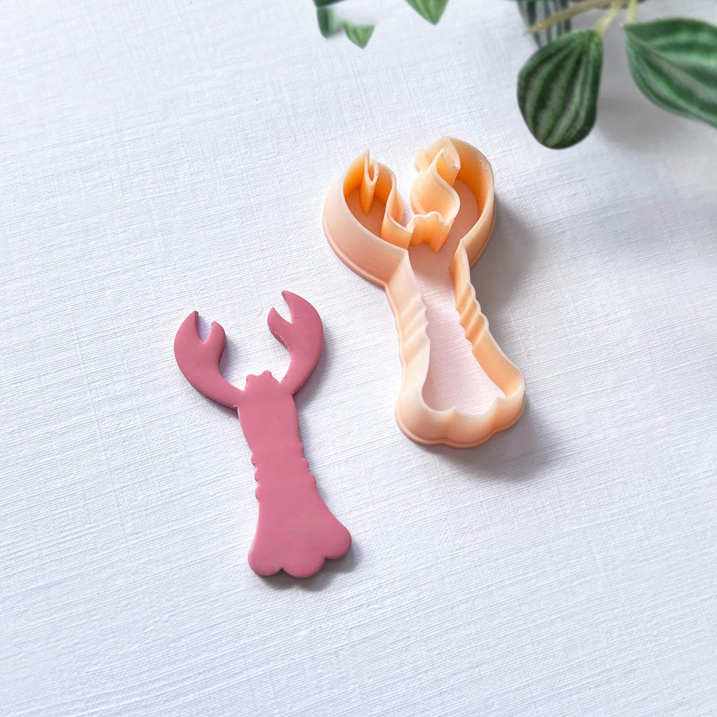 Pink polymer lobster cut out and the lobster shape clay cutter - on a light surface with green leaves in the background
