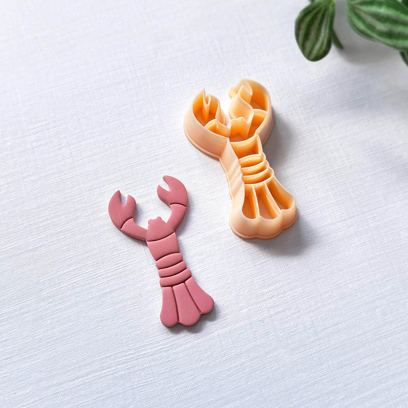 Peach lobster-shaped clay cutter and the pink polymer clay cut out on a light surface with a plant in the corner.