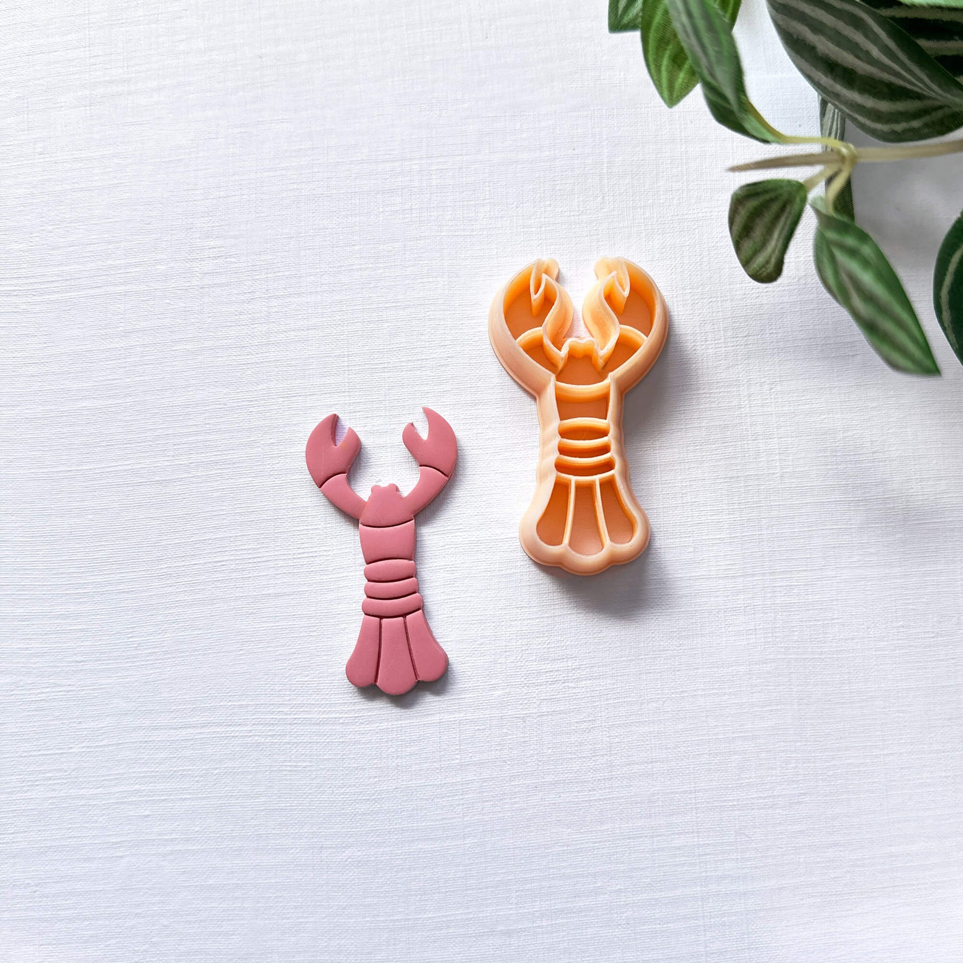 Peach lobster-shaped clay cutter and the pink polymer clay cut out on a light surface with a plant in the corner.