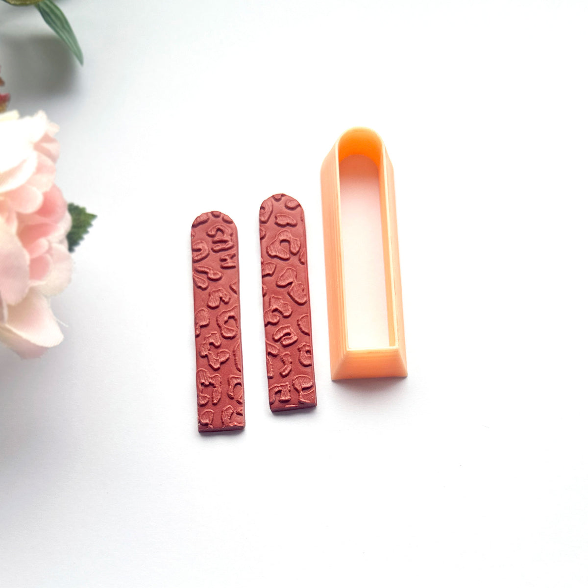 A peach-colored long arch clay cutter is shown next to brown clay cutouts, demonstrating shape on a clean white background with decorative green leaves.