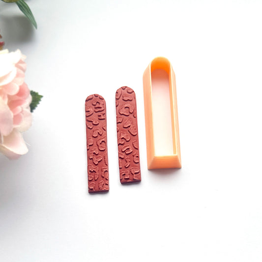 A peach-colored long arch clay cutter is shown next to brown clay cutouts, demonstrating shape on a clean white background with decorative green leaves.