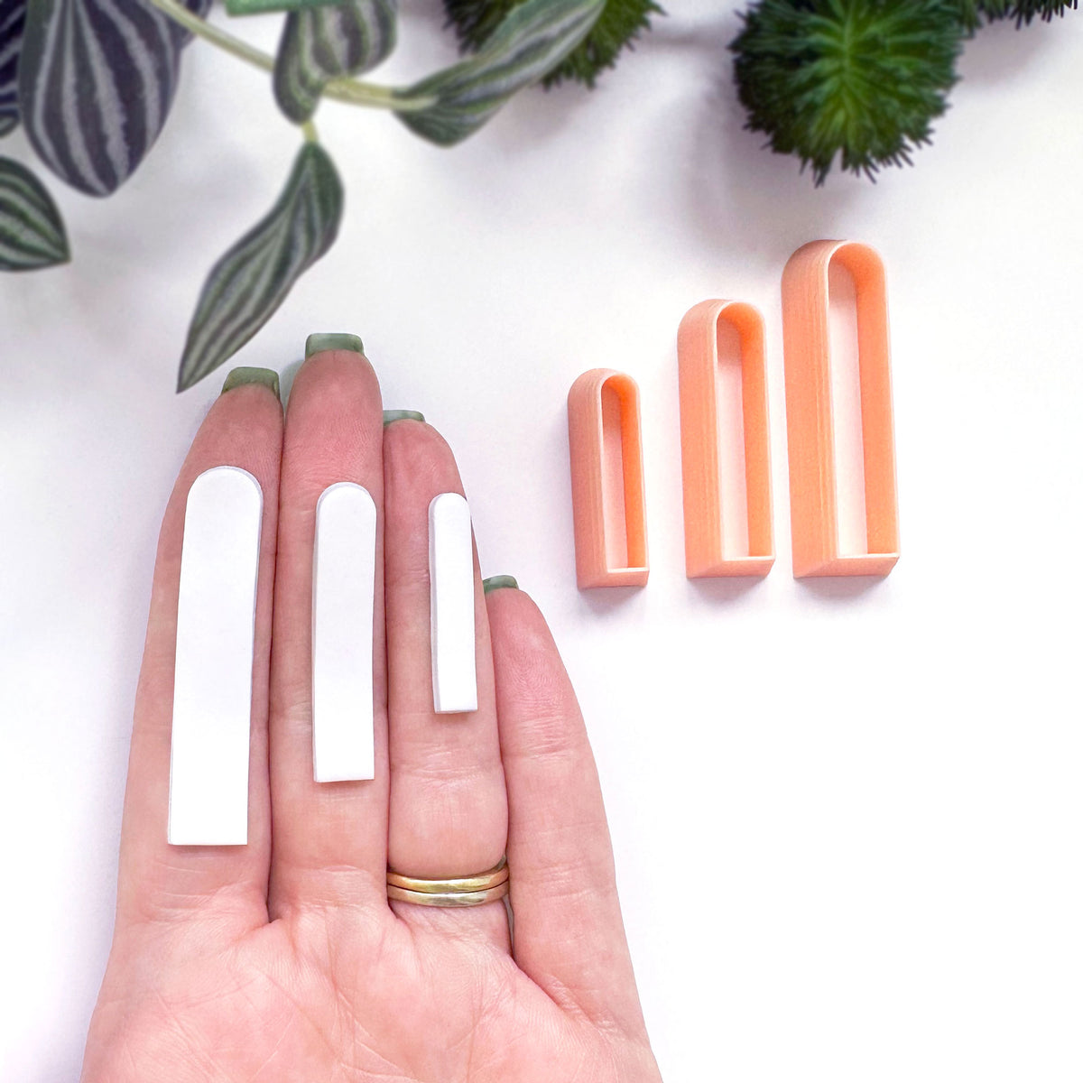Three peach-colored long arch clay cutters are shown next to a hand with white clay cutouts, demonstrating size progression on a clean white background with decorative green leaves.