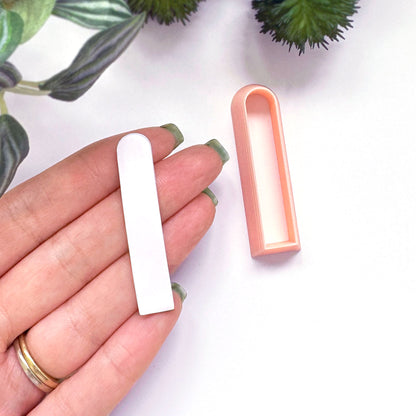 A hand with green nail polish holds a small white clay cut out next to a peach-colored plastic clay cutter mold  on a white background with decorative green leaves.