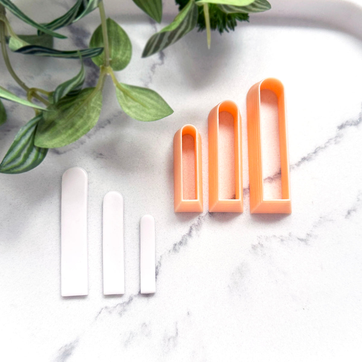 Three long slim arch clay cookie cutters in peach and three matching white clay cut outs arranged on a white marble surface with green leaves in the corner.