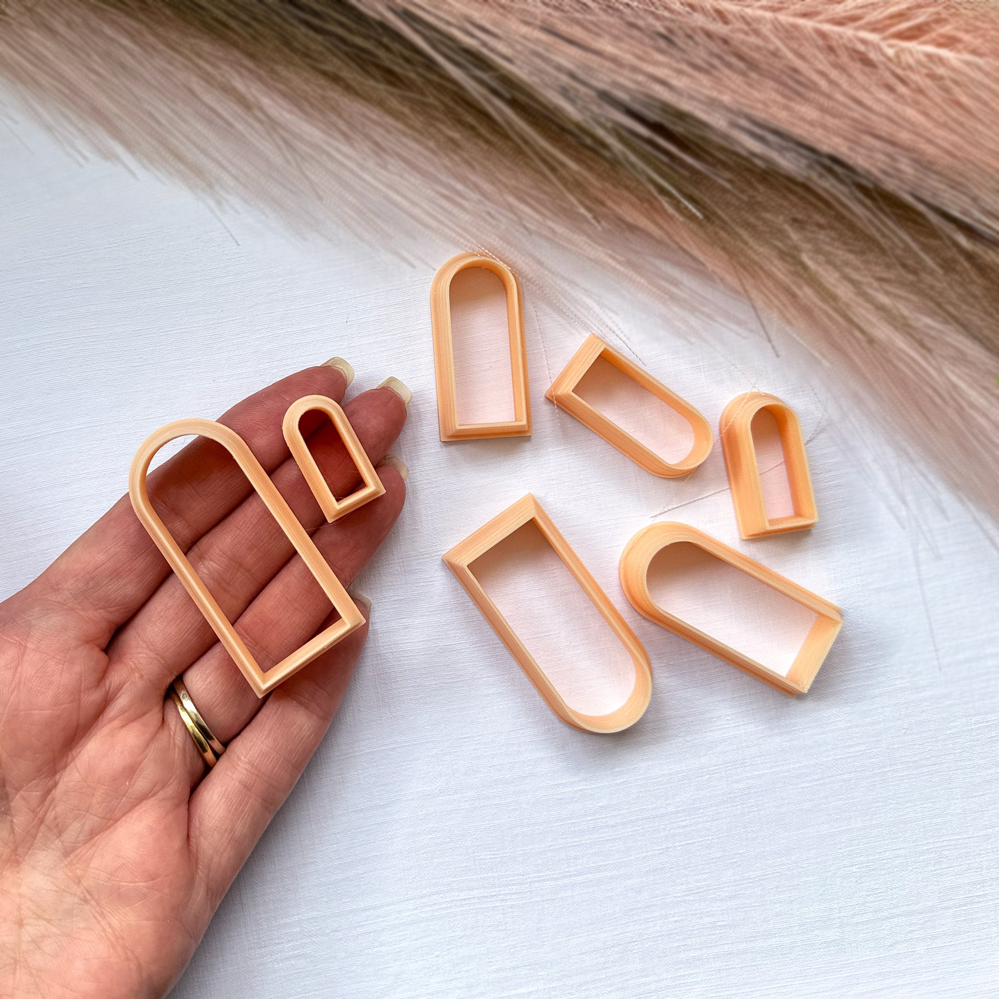 Set of peach-colored slim arch clay cutters on a white surface with a hand holding one clip.