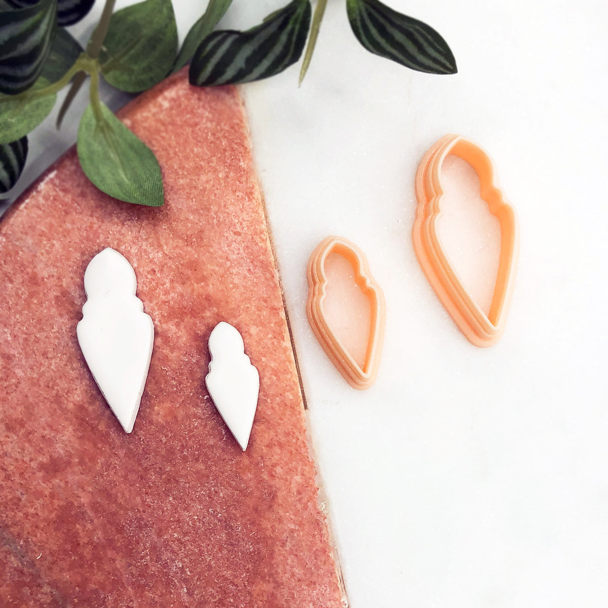 Two white 3D-printed white clay cut outs displayed on a textured pink surface next to two peach-colored plastic cookie cutters on a white marble background with green leaves.