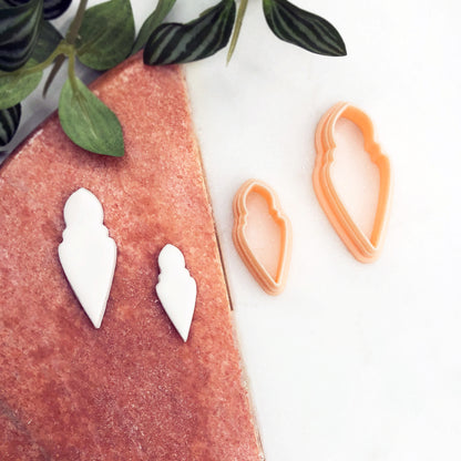 Two white 3D-printed white clay cut outs displayed on a textured pink surface next to two peach-colored plastic cookie cutters on a white marble background with green leaves.