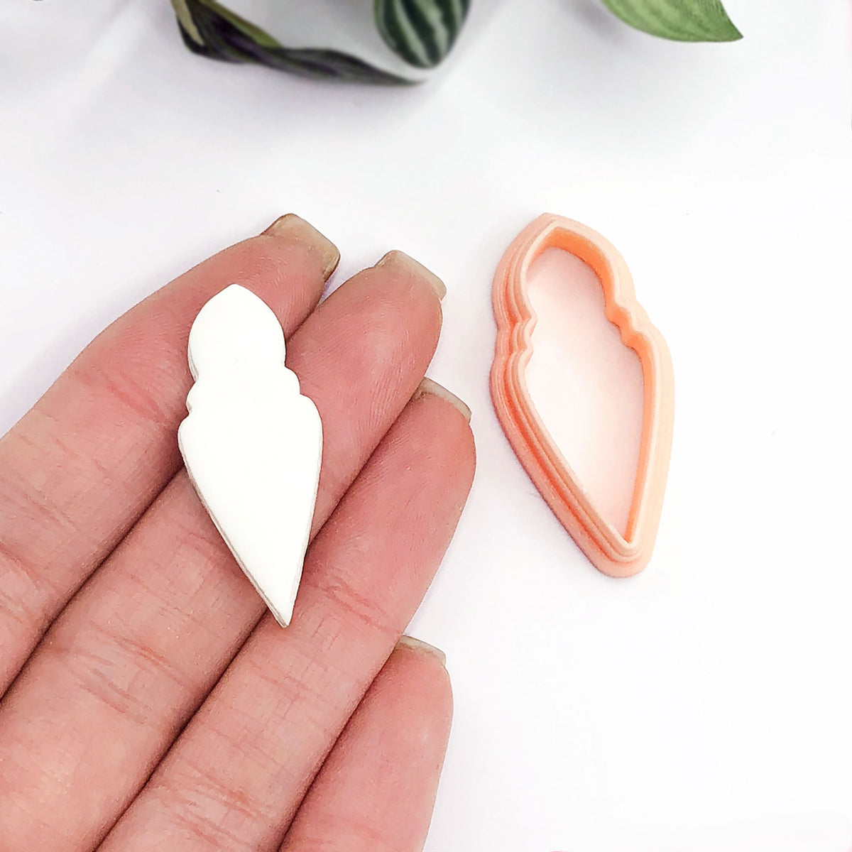 A hand holds a small white long bauble-shaped clay cutout next to a peach-colored 3d printed cookie cutter on a clean white background.