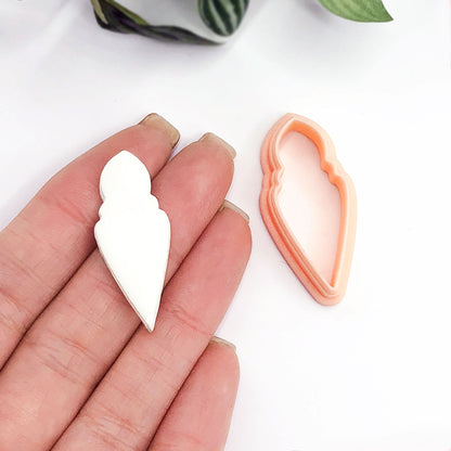 A hand holds a small white long bauble-shaped clay cutout next to a peach-colored 3d printed cookie cutter on a clean white background.