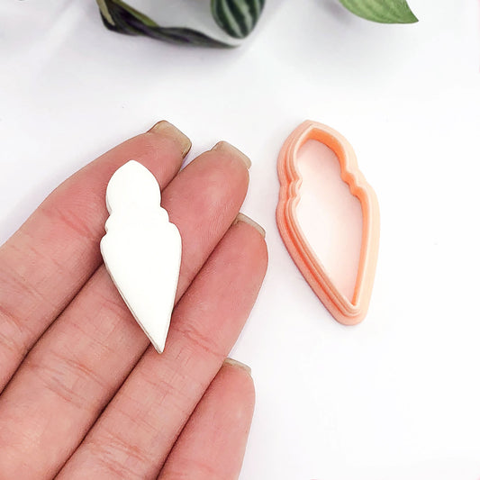 A hand holds a small white long bauble-shaped clay cutout next to a peach-colored 3d printed cookie cutter on a clean white background.