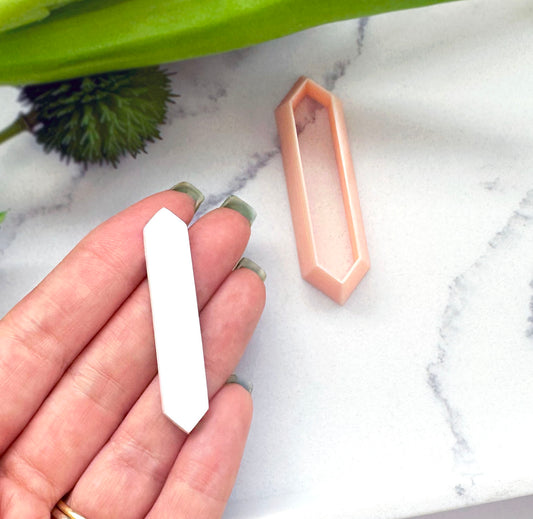 A hand holds a white hexagon crystal-shaped polymer clay cut out piece next to a peach-colored clay cutter on a white marble surface.