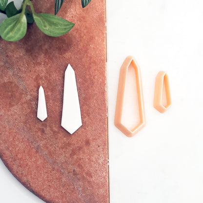 Two white clay cut outs and two pointed geometric cookie cutters in peach are displayed on a split white and terracotta background with green leaves in the corner.