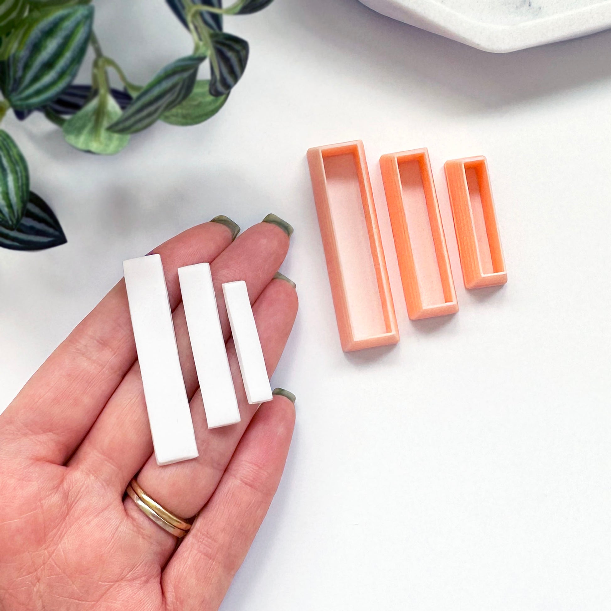 A hand holds three small white rectangular Polymer clay bars, with a peach-colored rectangular clay cutters beside it on a white surface.