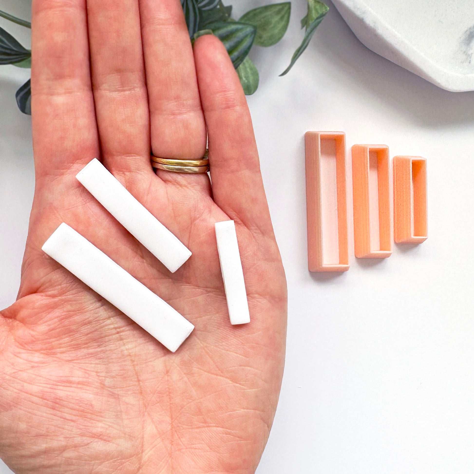 A hand holds three small white rectangular Polymer clay bars, with peach-colored rectangular clay cutters beside it on a white surface.