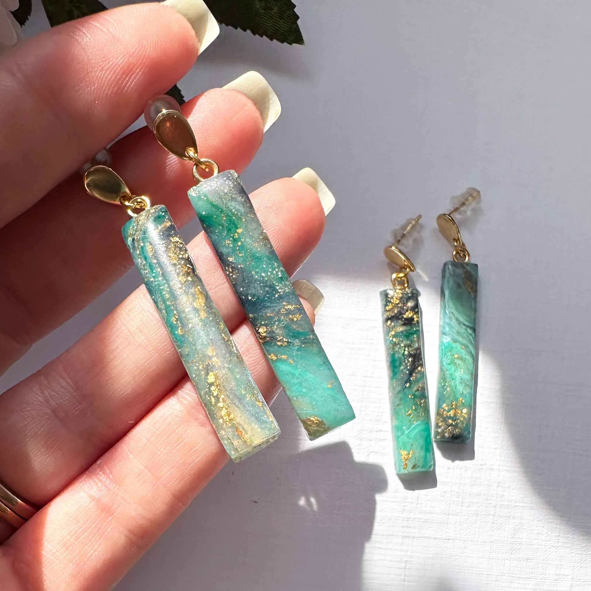 A pair of dangling earrings with rectangular marbled green and white polymer clay pendants and gold-tone posts, shown being held over a white surface with the matching pair placed beside it.