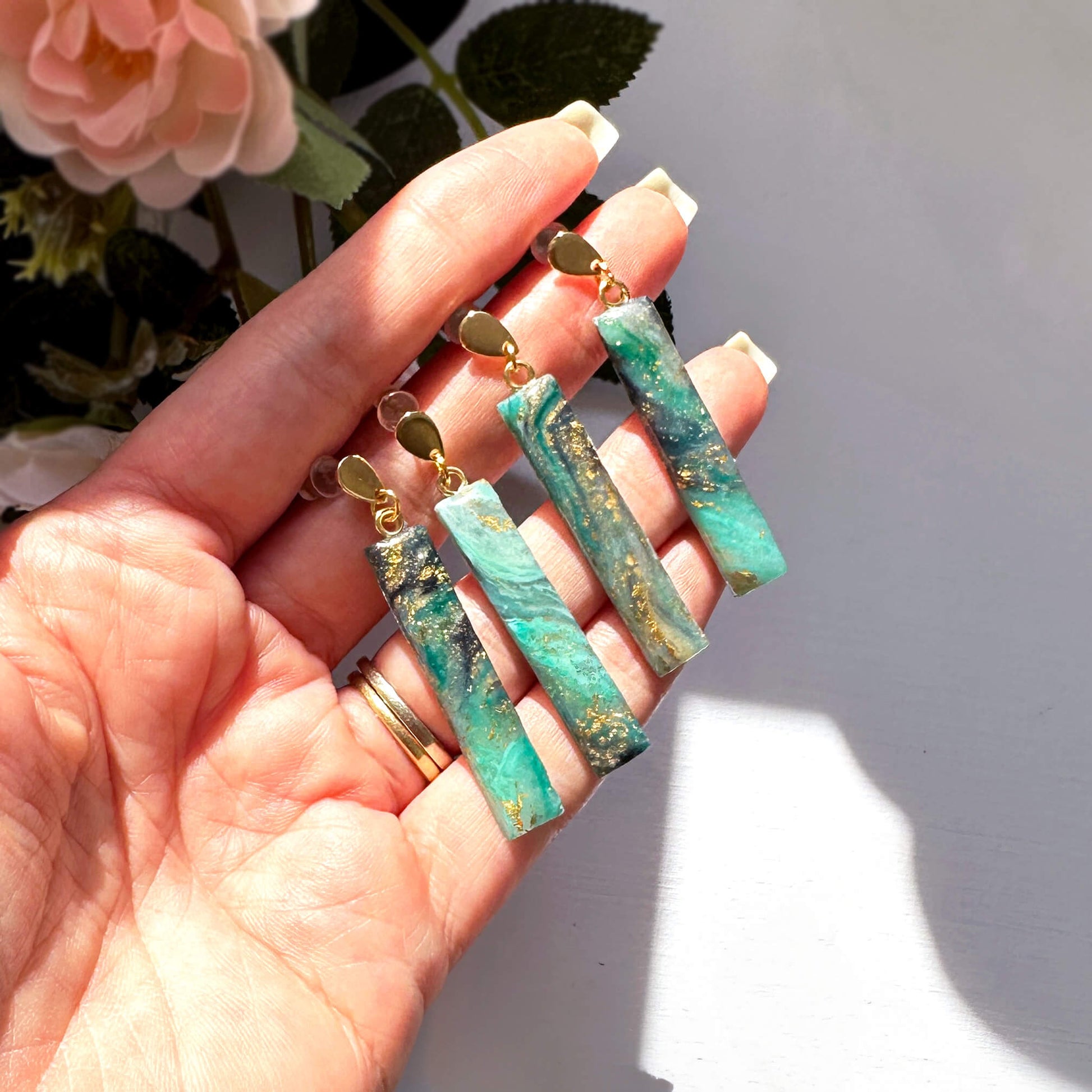 A hand holding four rectangular drop earrings with marbled turquoise-green stone and gold flecks, attached to gold-tone posts. Pink flowers and green leaves are softly blurred in the background against a bright white surface.