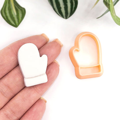 A white glove-shaped item held in a person's hand next to an orange glove-shaped cookie cutter on a white background with green plant leaves in the corner.