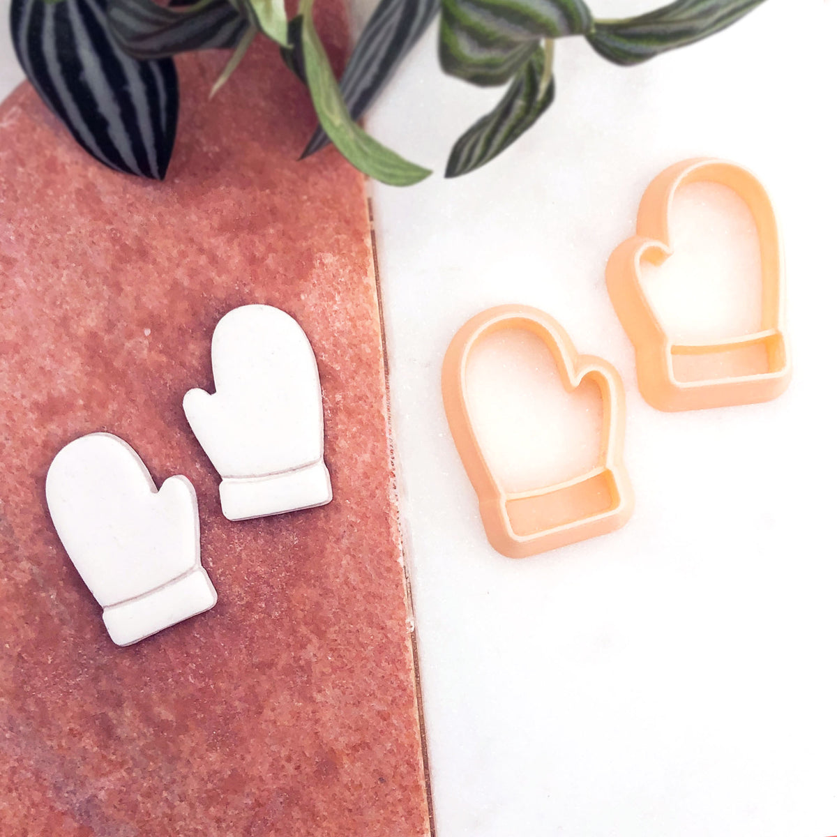A mirror set of small white mitten-shaped clay cut outs, with two peach-colored mitten-shaped cookie cutters placed on a white surface. A green plant leafy branch is visible in the upper right corner.