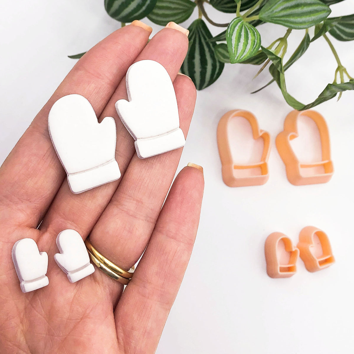 A hand holds 2 mirror sets of small white mitten-shaped clay cut outs, with two sets of peach-colored mitten-shaped cookie cutters placed on a white surface nearby. A green plant leafy branch is visible in the upper right corner.