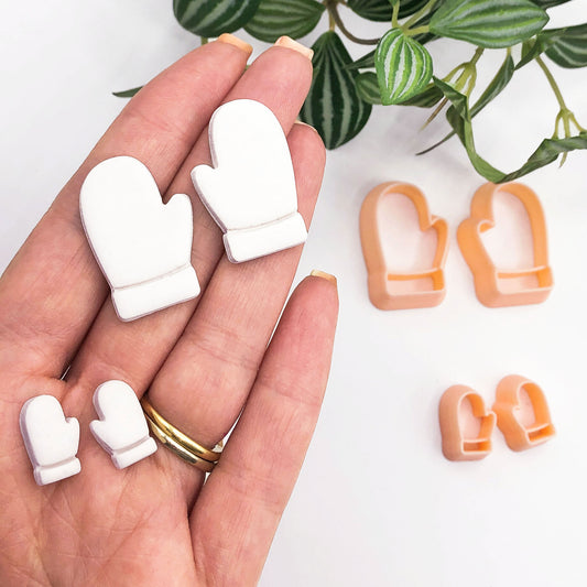 A hand holds 2 mirror sets of small white mitten-shaped clay cut outs, with two sets of peach-colored mitten-shaped cookie cutters placed on a white surface nearby. A green plant leafy branch is visible in the upper right corner.
