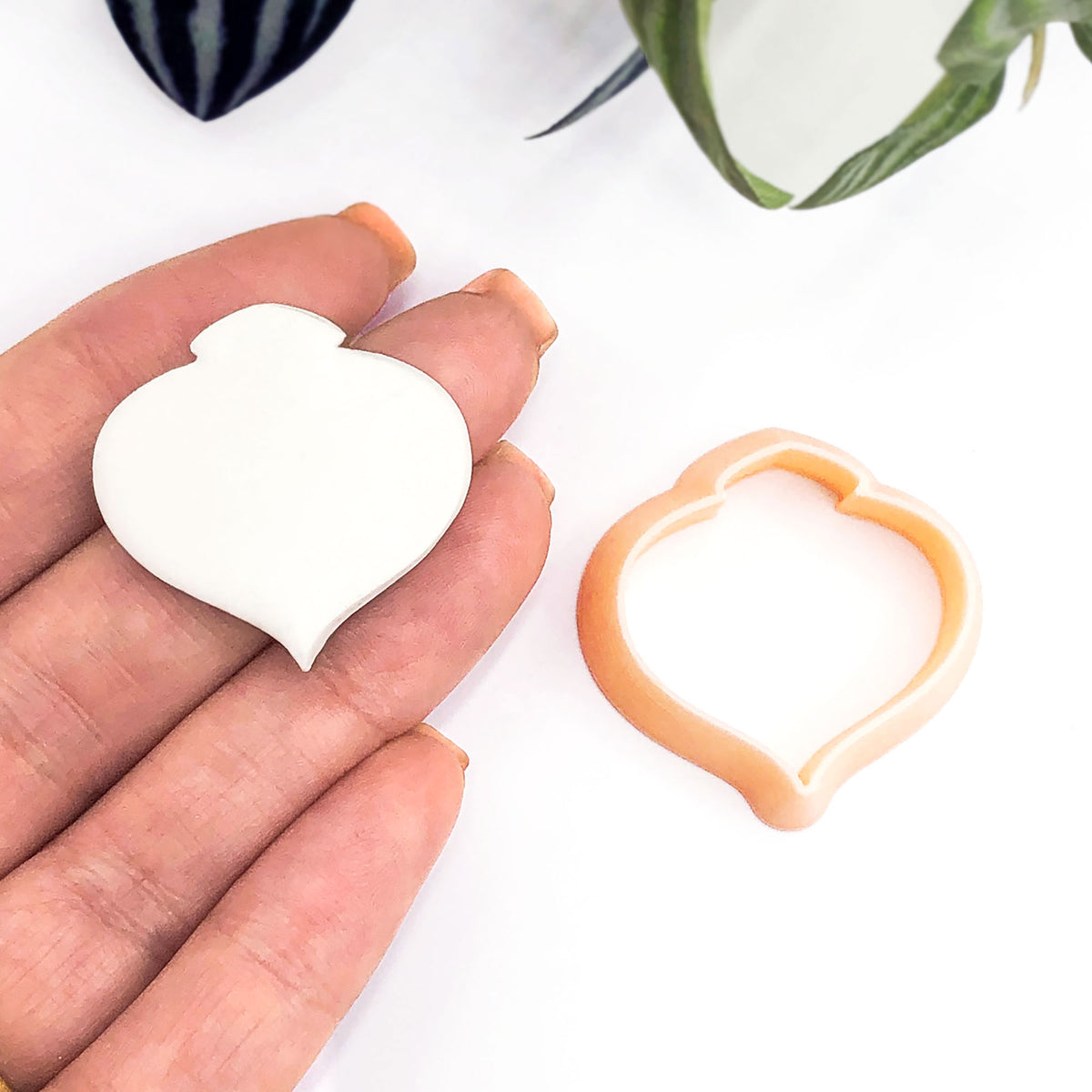 A hand holds a small white moroccan bauble-shaped clay piece next to a peach-colored clay cookie cutter on a white background with blurred green leaves.