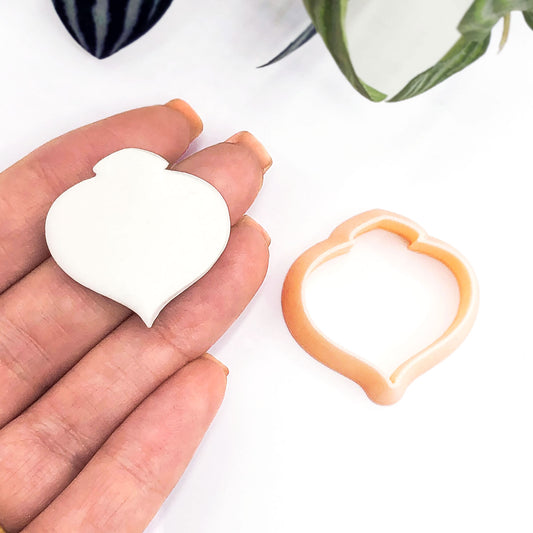 A hand holds a small white moroccan bauble-shaped clay piece next to a peach-colored clay cookie cutter on a white background with blurred green leaves.