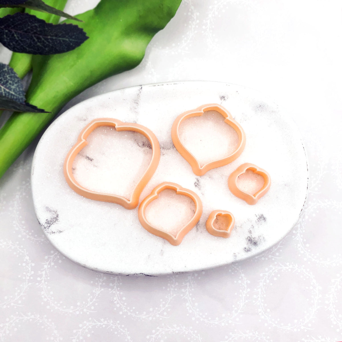 Four peach-colored bauble cookie cutters in different sizes arranged on a white marble tray, with green leaves in the background.