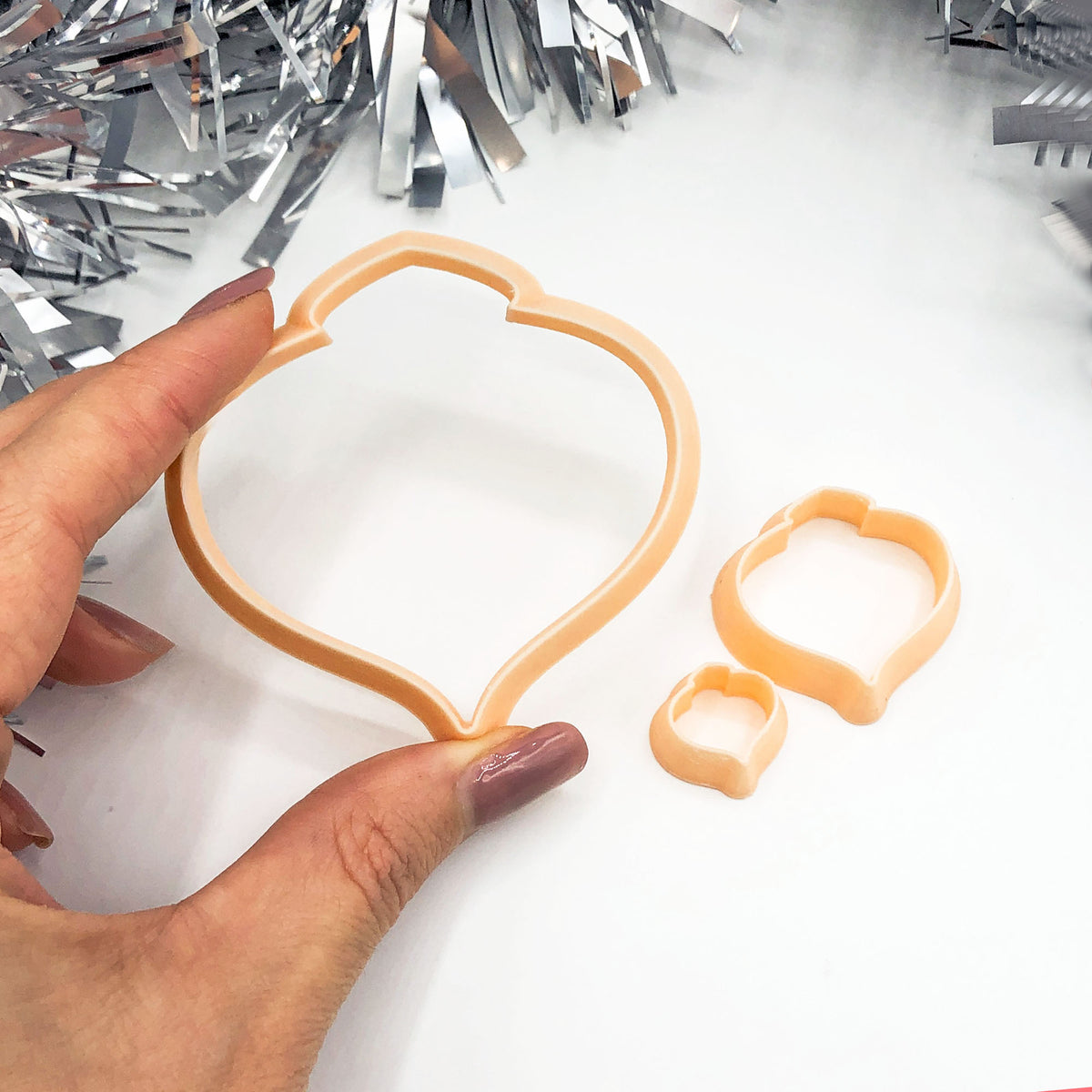 A peach-colored plastic cookie cutter set consisting of three nested shapes, held by a hand over a white background with silver tinsel decorations.